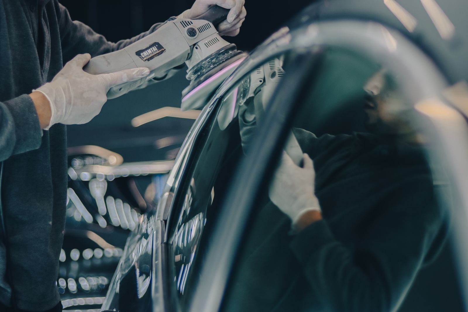 Gloved hands running a dual-action polisher along the glossy dark bodywork of a car in a dim detailing studio