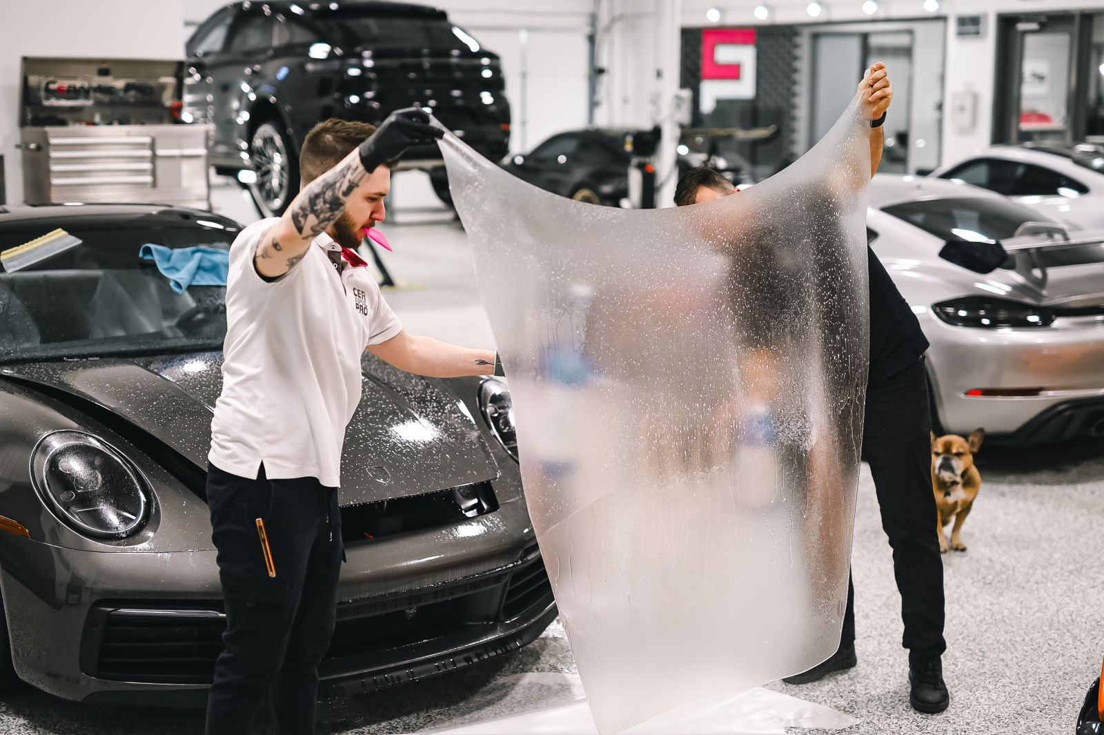 Two detailers holding up a large sheet of clear paint protection film above the wet bonnet of a grey Porsche 911 in a detailing studio