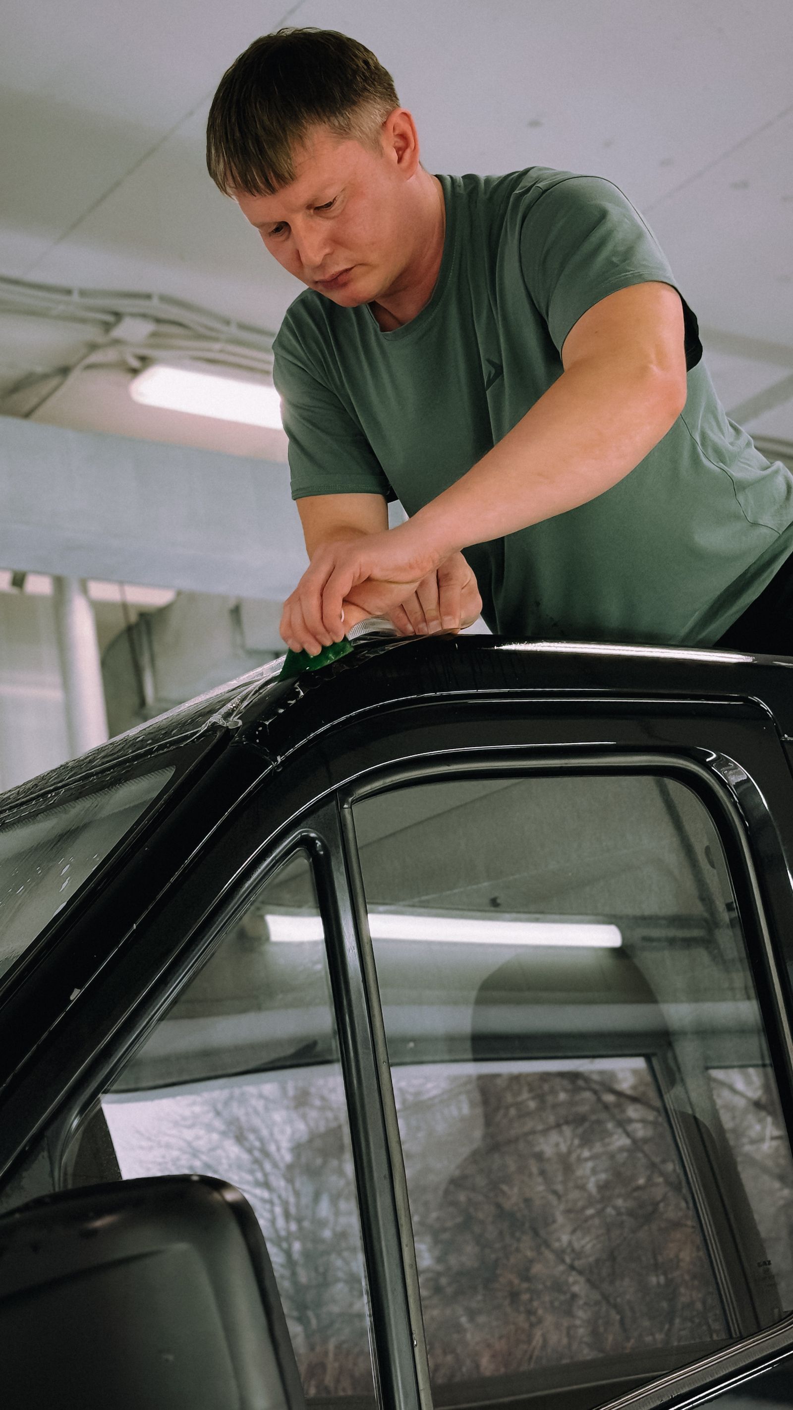 Technician squeegeeing wet window tint film onto the glass of a black car inside a garage