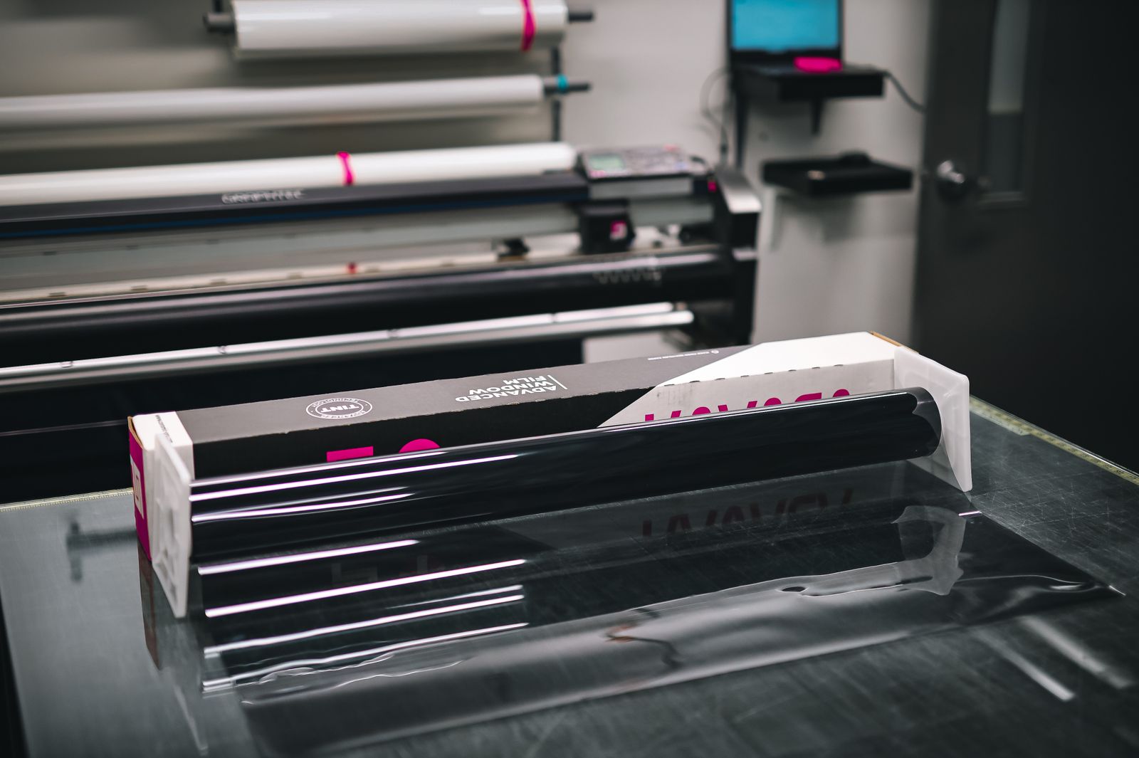 Roll of gloss black film unrolled on a wrap shop cutting table in front of a plotter loaded with more film rolls