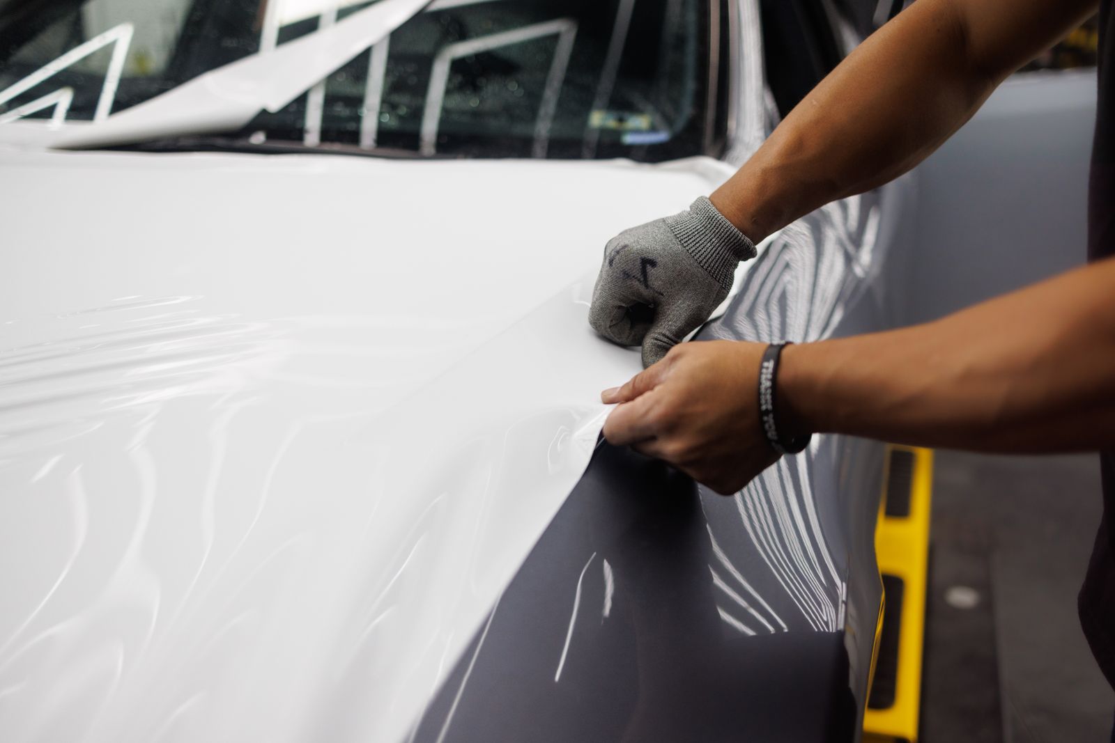 Installer's gloved hands laying a large sheet of gloss white vinyl wrap film across the bonnet and wing of a dark grey car in a wrap bay