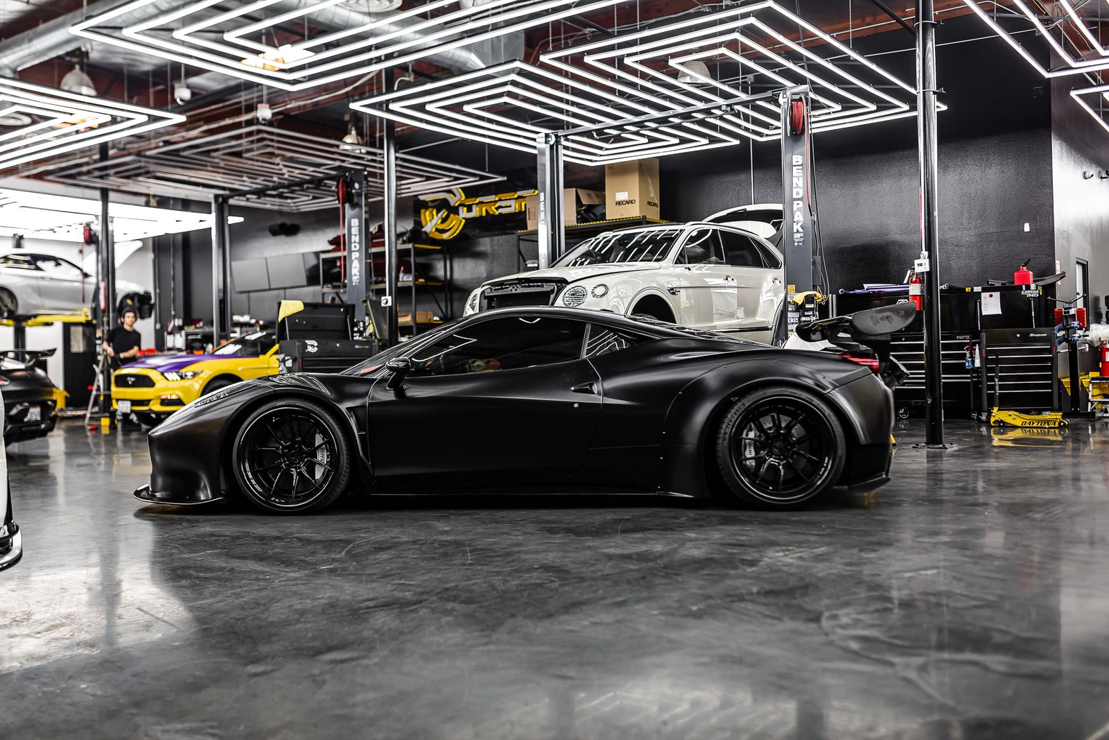 Matte black widebody Ferrari on the floor of a high-end wrap workshop, flanked by two-post lifts, rolling tool chests and LED ceiling lighting