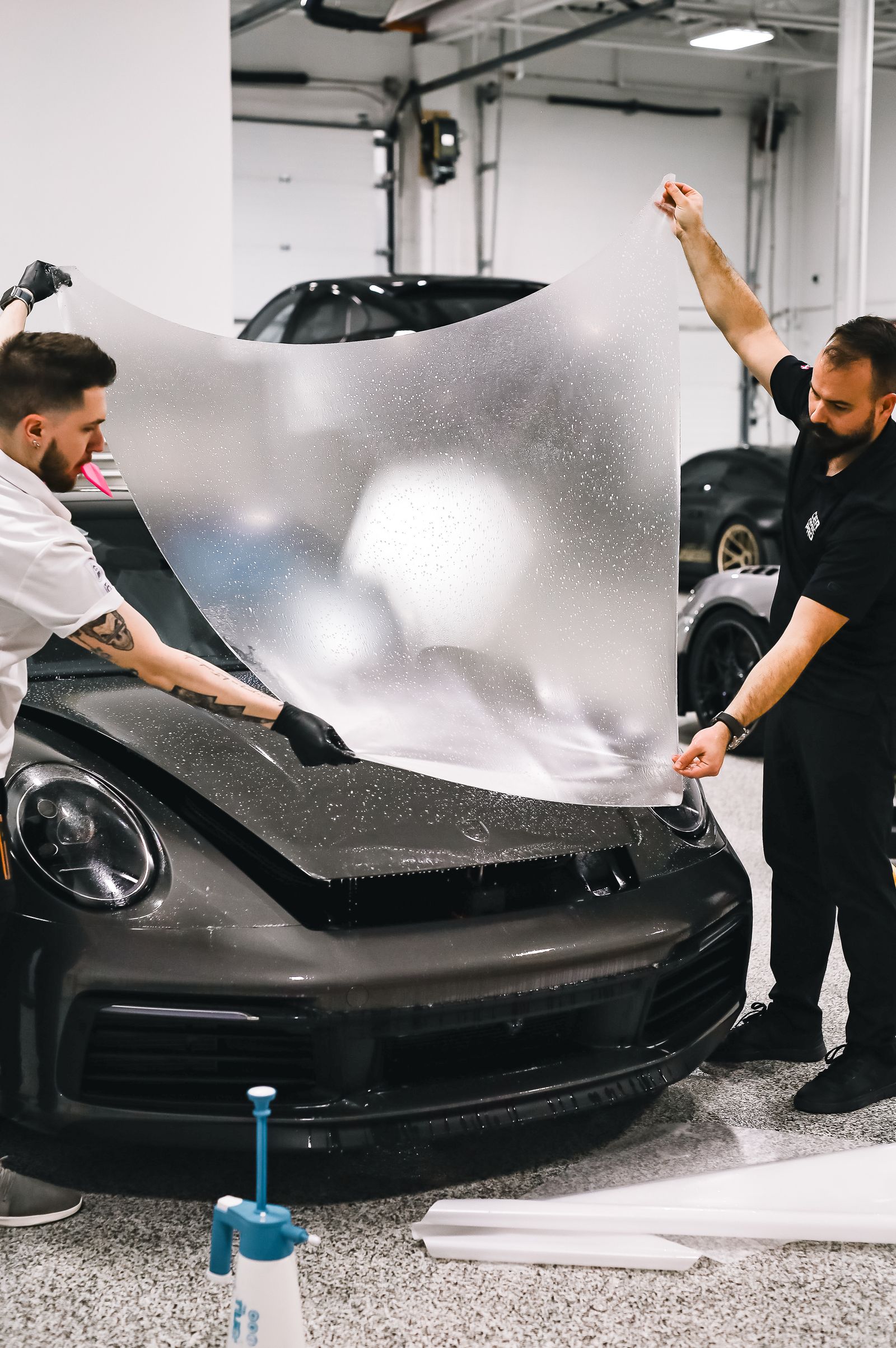 Two installers stretching a large sheet of transparent protection film over the bonnet of a grey Porsche 911 in a workshop