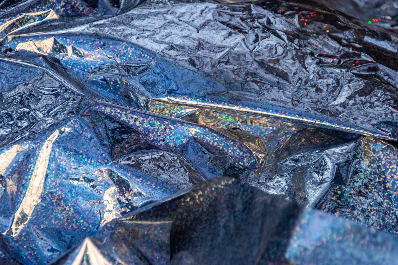 Macro of crumpled holographic chrome film, faceted creases shifting between silver, blue and violet with iridescent glitter flecks