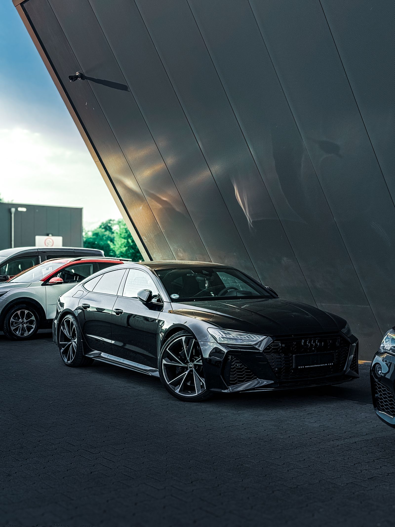 Gloss black Audi RS7 Sportback beneath a dramatic angled metal facade, shot from a low front three-quarter angle