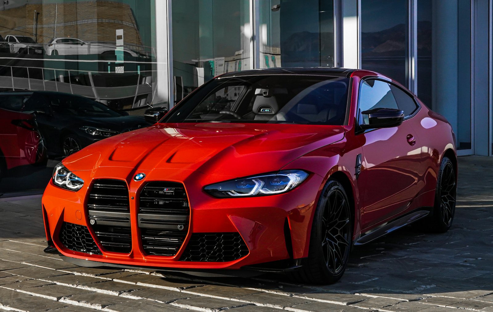 Glossy red BMW M4 Competition with the large vertical kidney grille, parked on stone pavers against dark glass