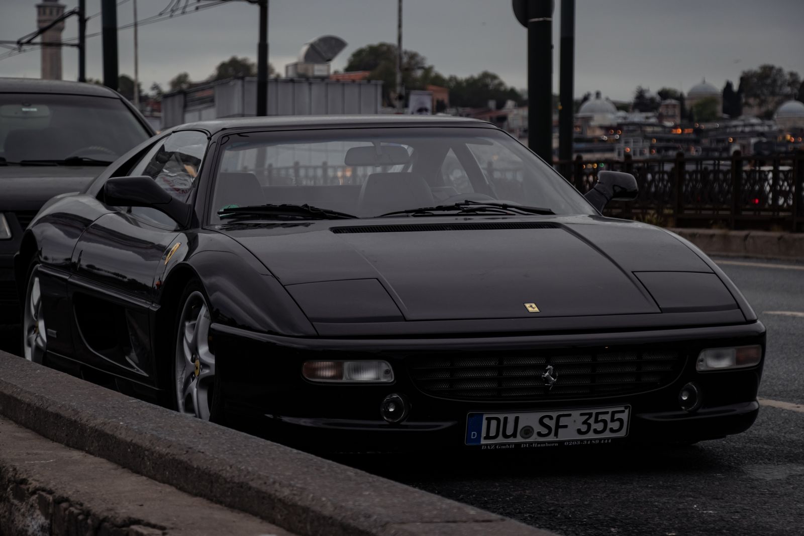 Gloss black Ferrari F355 on an Istanbul bridge at dusk, city skyline dissolving into haze behind it