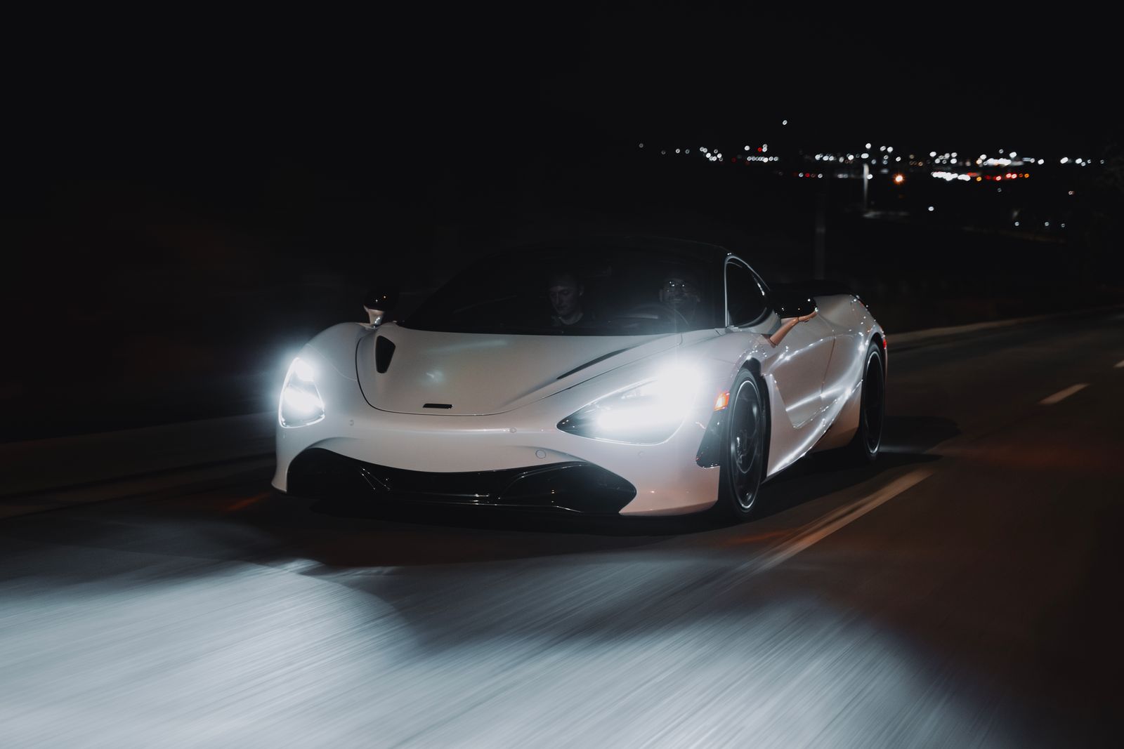 White McLaren 720S at speed on a dark road at night, headlights blazing and the road blurred beneath it