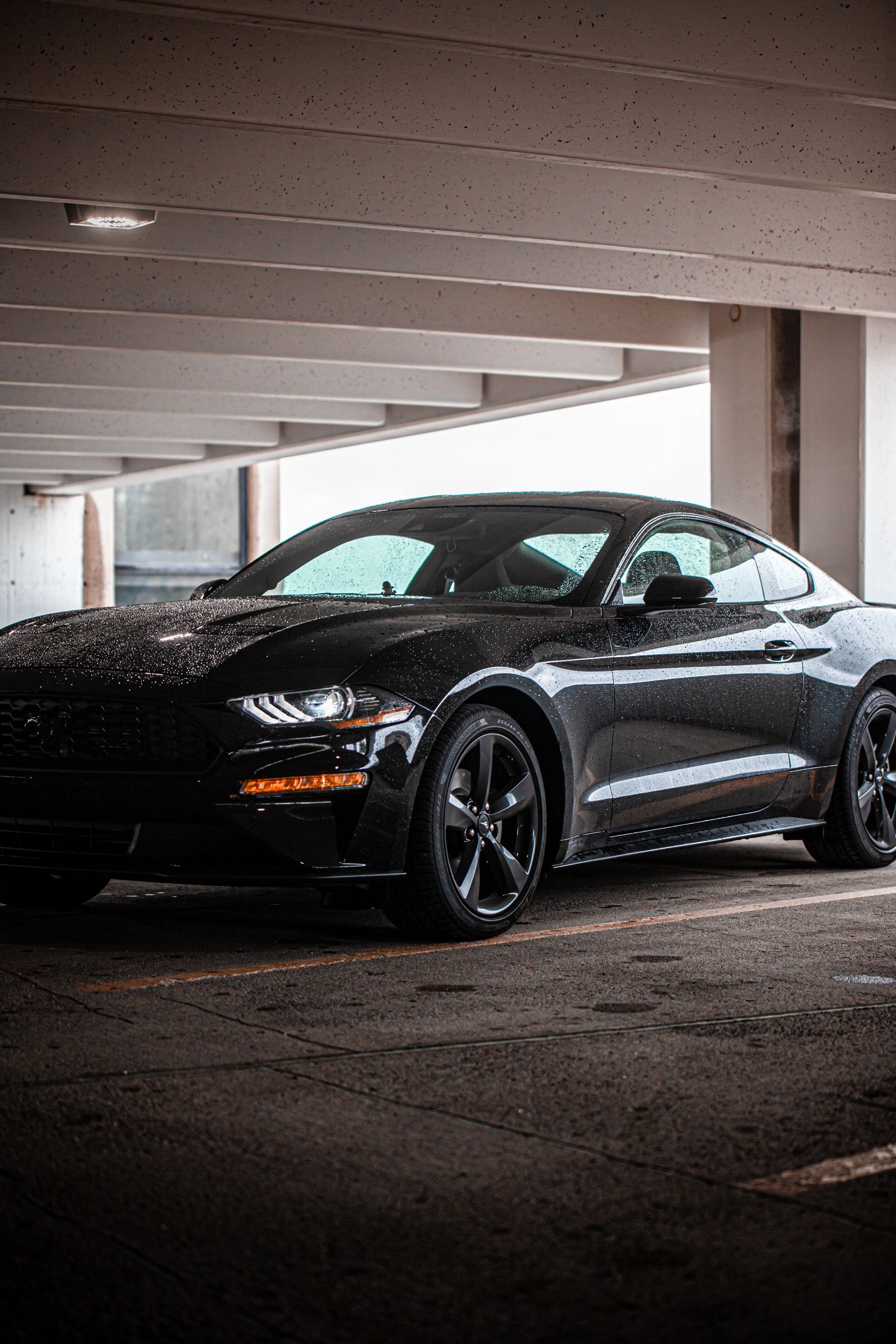 Gloss black Ford Mustang beaded with rain, parked in a dim concrete parking garage