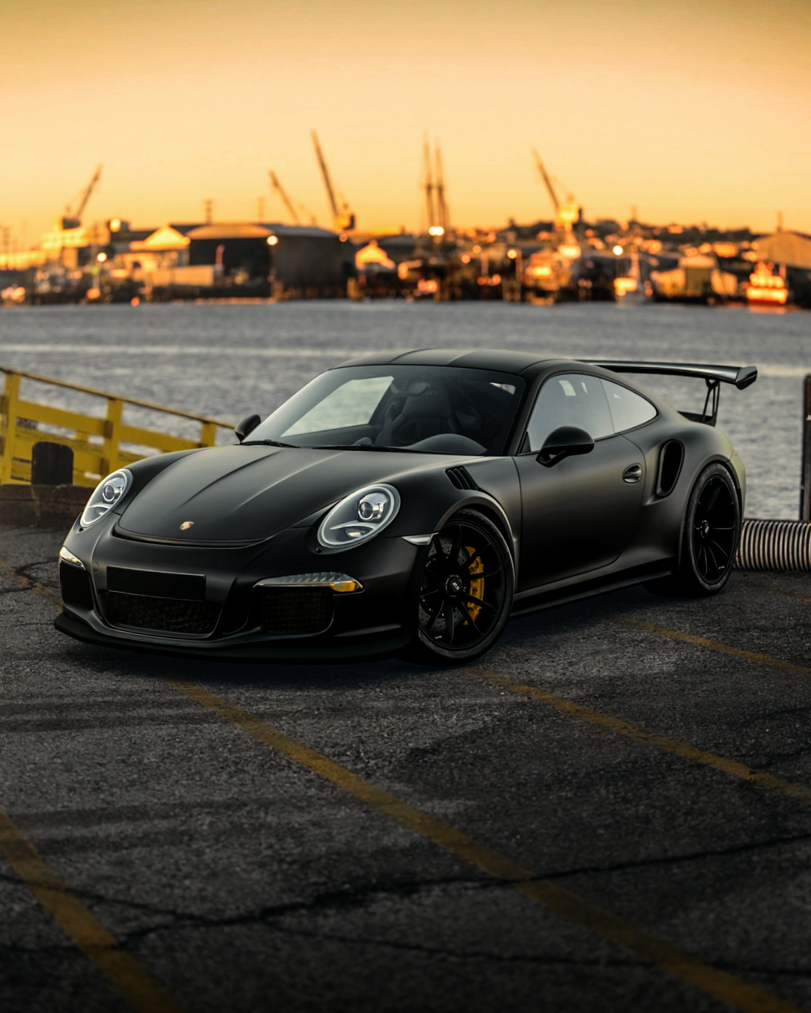 Satin black Porsche 911 GT3 RS parked on a harbour dock at sunset, rear wing raised against silhouetted cranes